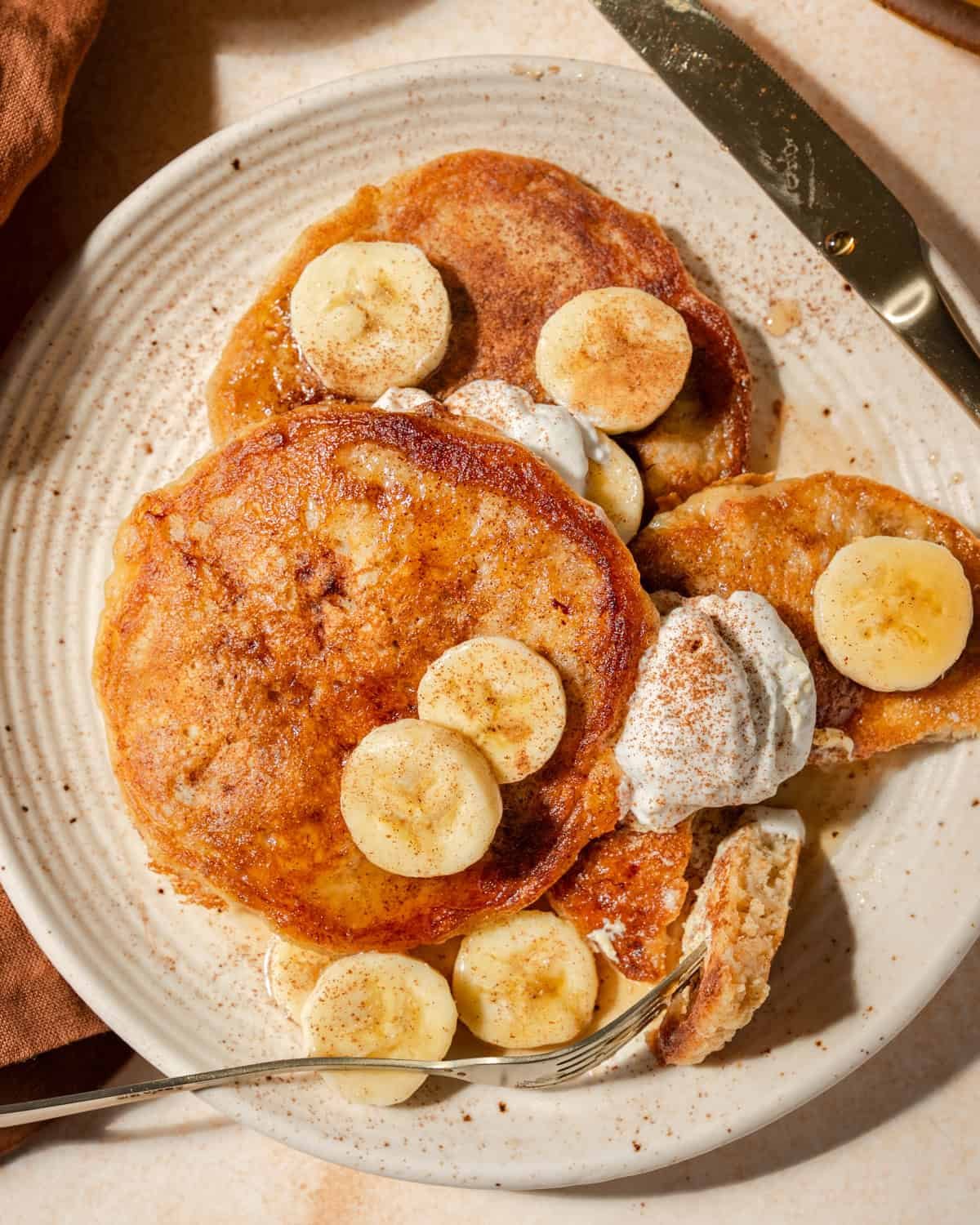 Close up overhead photo of person taking a bite from 3 banana pancakes on a plate.
