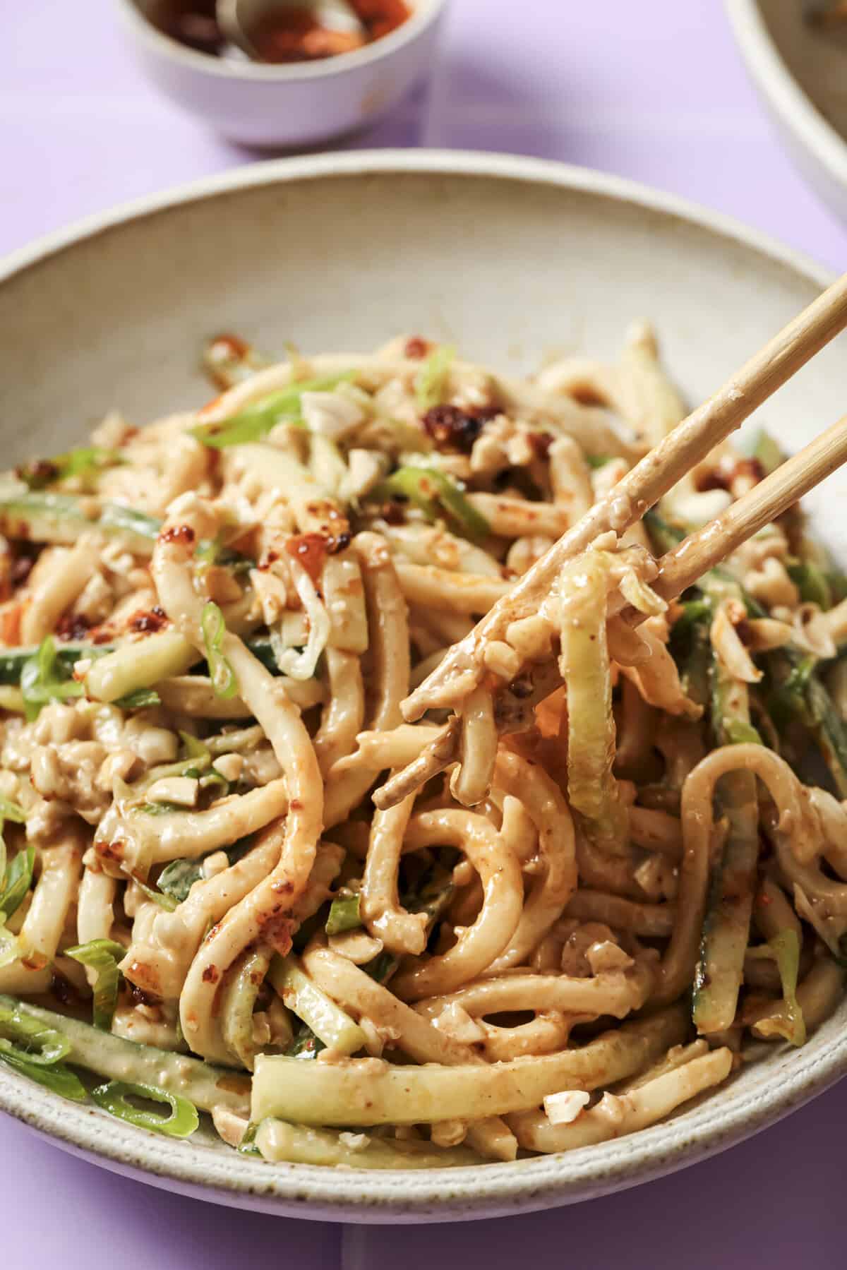 Close-up of peanut noodles with cucumber, green onions, and chili oil, held with chopsticks in a light bowl.