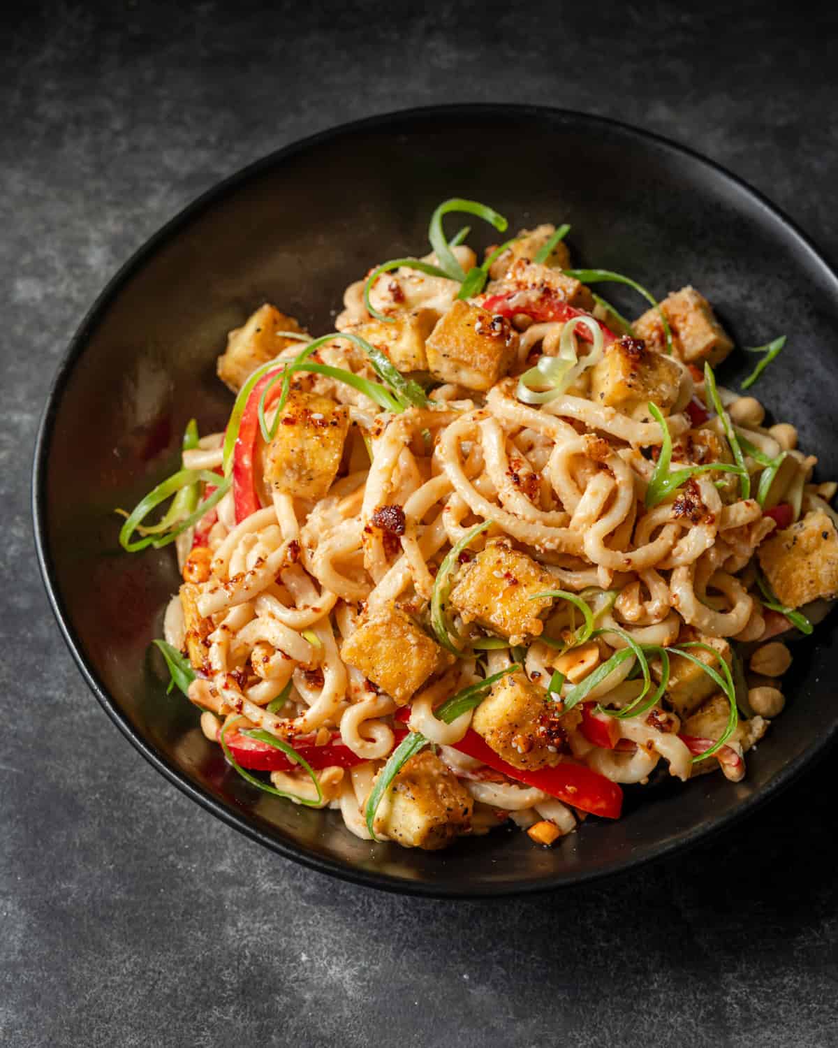 Overhead view of peanut noodles with tofu, red peppers, green onions, and chili oil.