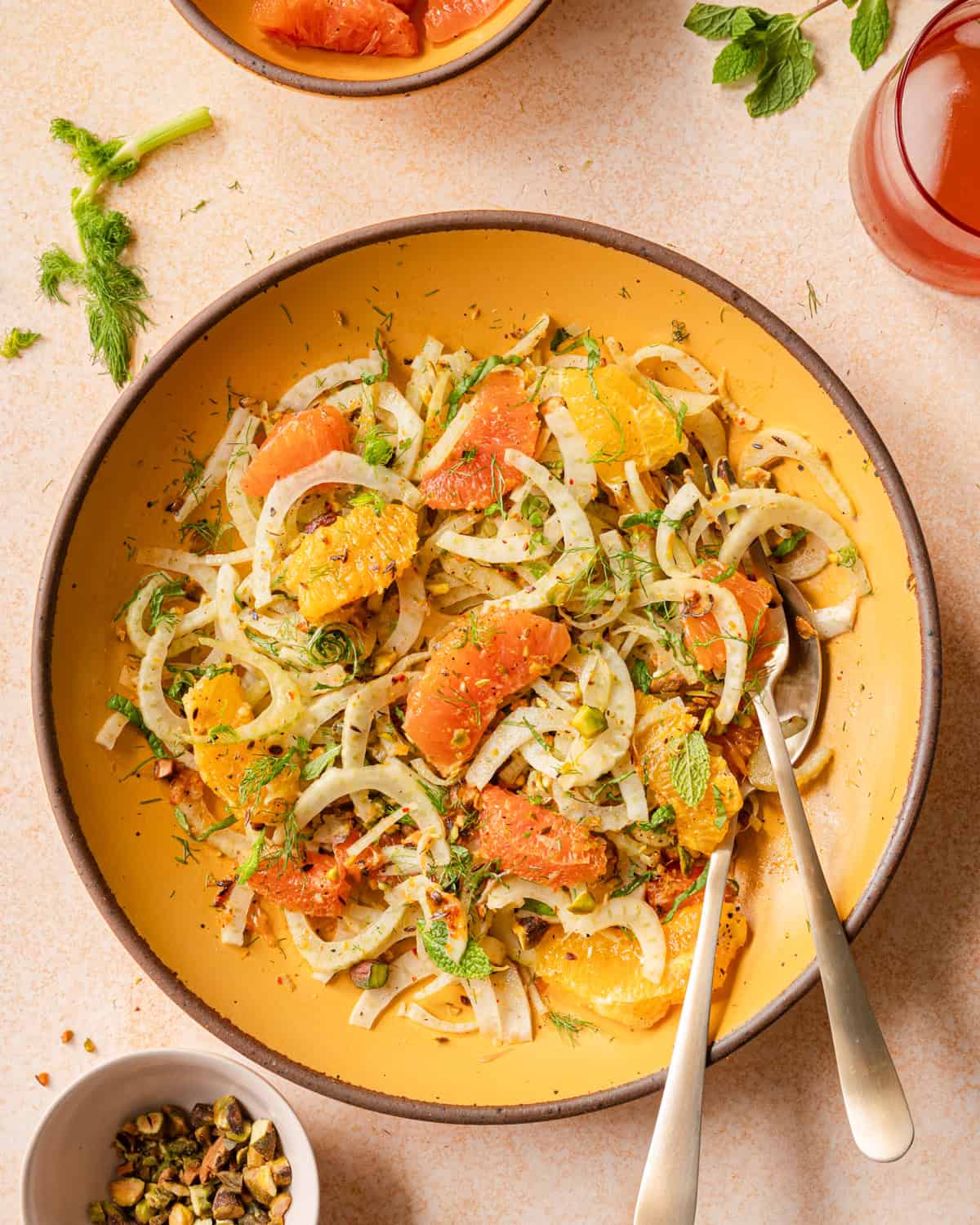 fennel orange salad in a yellow bowl with gold silverware on a pink surface.
