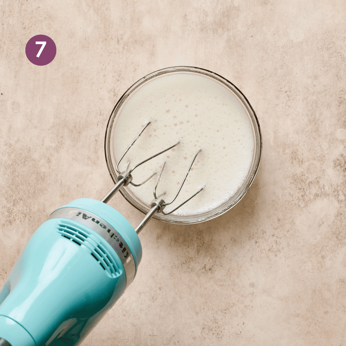 light blue electric mixer in a glass bowl of whipped aquafaba on a light pink surface.