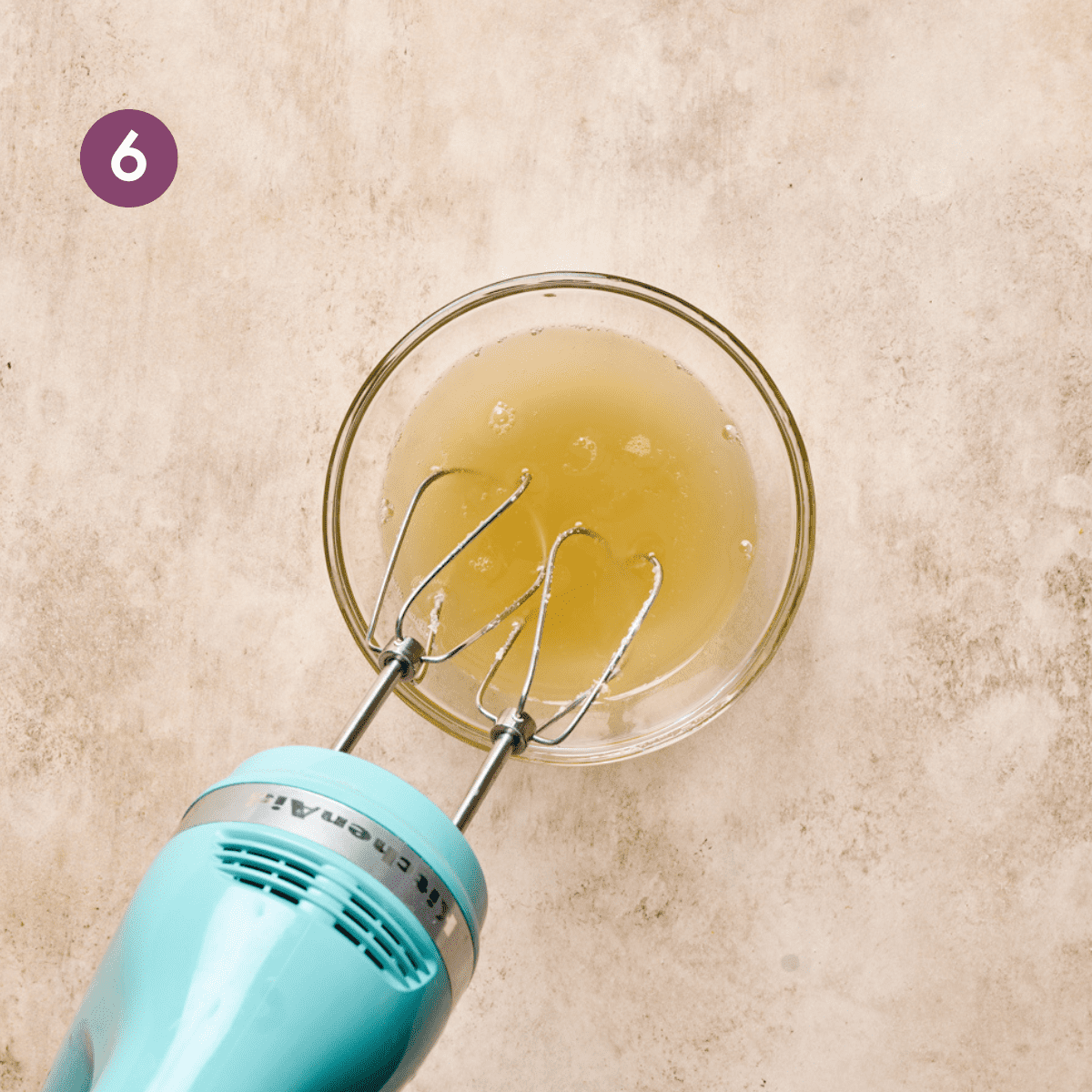 light blue electric mixer in a glass bowl of aquafaba on a light pink surface.