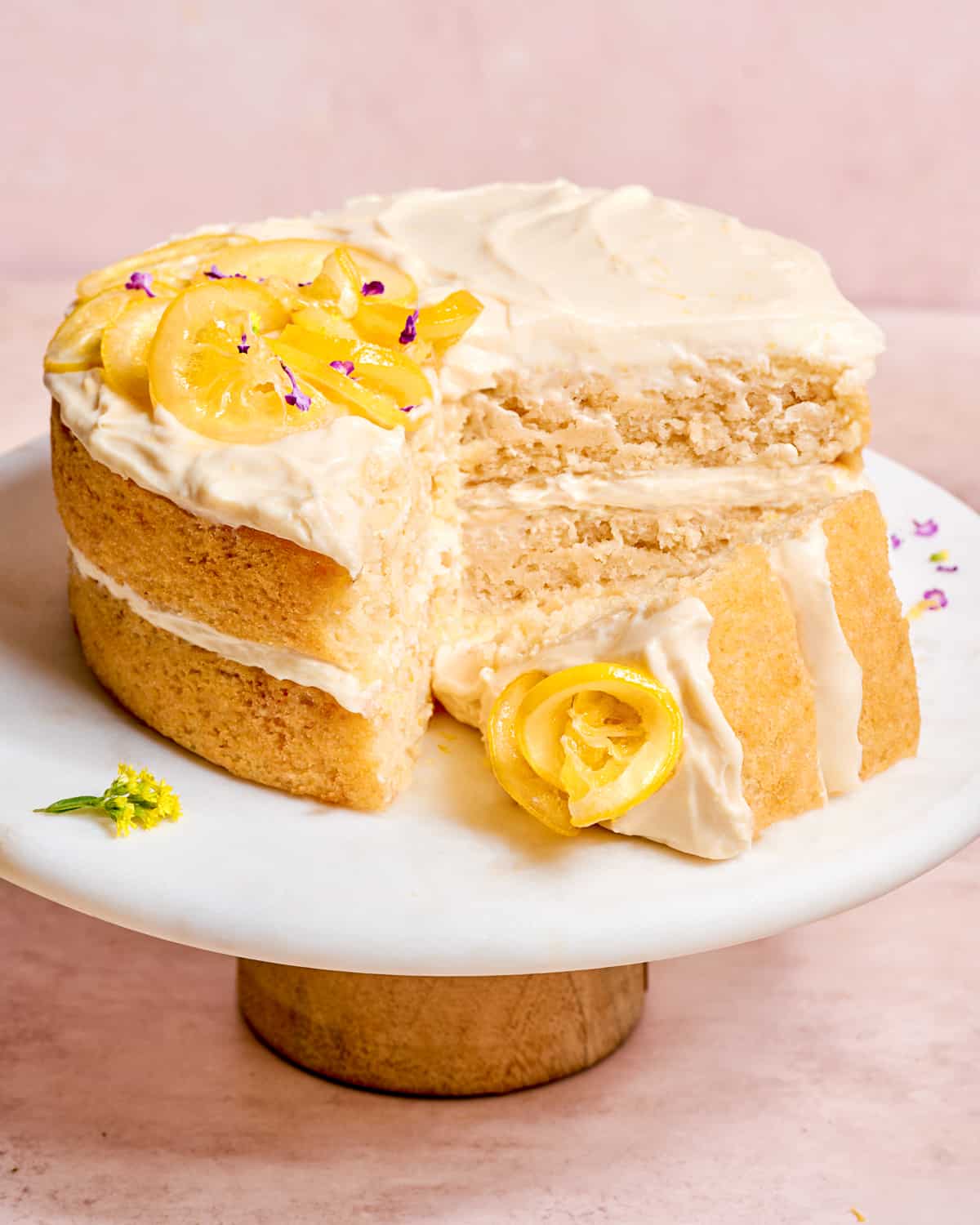 vegan lemon cake with cream cheese frosting on a white cake stand, with one slice out on its side.