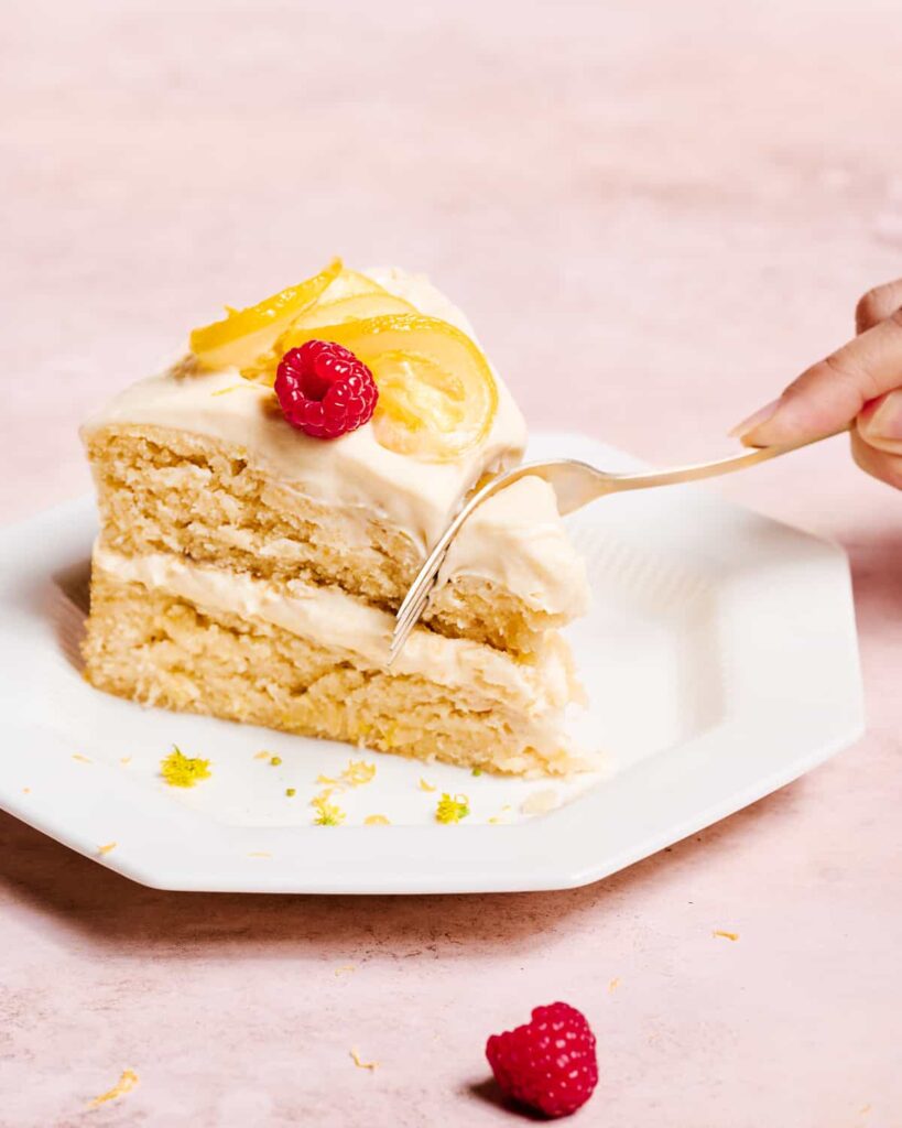 two-layer vegan lemon cake with lemon cream cheese frosting on a white cake stand topped with candied lemons.